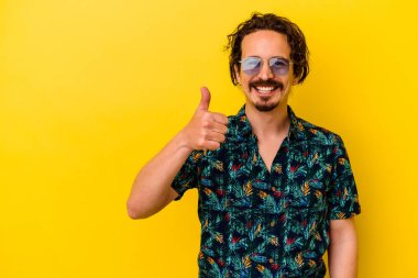 Sarı arka planda izole edilmiş yaz kıyafetleri giyen genç beyaz adam gülümsüyor ve başparmağını kaldırıyor.