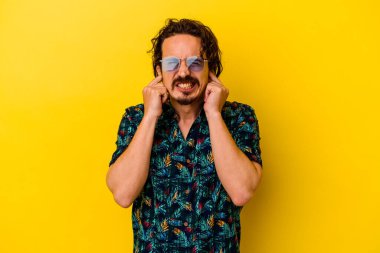 Sarı arka planda izole edilmiş yaz kıyafetleri giyen genç beyaz adam elleriyle kulaklarını kapatıyor..
