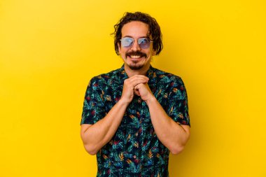 Sarı arka planda izole edilmiş yaz kıyafetleri giyen genç beyaz adam ellerini çenesinin altında tutuyor ve mutlu bir şekilde yan tarafa bakıyor..