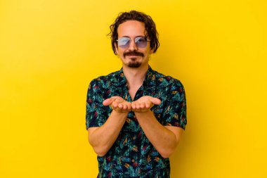 Sarı arka planda izole edilmiş yaz kıyafetleri giyen genç beyaz bir adam elinde avuç içi bir şeyle kameraya sunuyor..