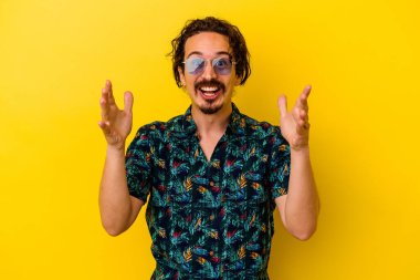Sarı arka planda izole edilmiş yaz kıyafetleri giyen genç beyaz adam hoş bir sürpriz alıyor, heyecanlı ve el kaldırıyor..