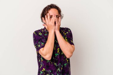 Beyaz arka planda izole edilmiş genç beyaz bir adam kameraya parmaklarıyla göz kırpıyor, yüzünü kapatmaktan utanıyor..