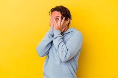 Sarı arka planda izole edilmiş genç beyaz adam parmaklarının arasından korkmuş ve gergin bir şekilde göz kırp..