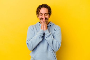 Sarı arka planda izole edilmiş genç beyaz bir adam el ele tutuşup ağız kenarında dua ediyor, kendinden emin hissediyor..