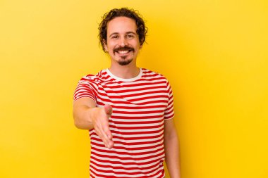 Sarı arka planda izole edilmiş genç beyaz adam selamlama jesti olarak kameraya elini uzatıyor..