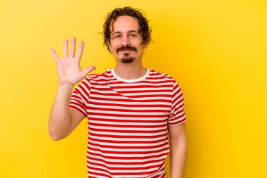 Sarı arka planda izole edilmiş genç beyaz adam gülümseyerek beş numarayı parmaklarıyla gösteriyor..