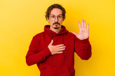 Sarı arka planda izole edilmiş genç beyaz adam yemin ediyor, elini göğsüne koyuyor..