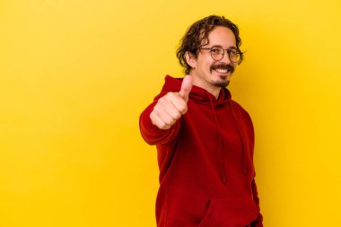 Sarı arka planda izole edilmiş genç beyaz adam gülümsüyor ve başparmağını kaldırıyor.