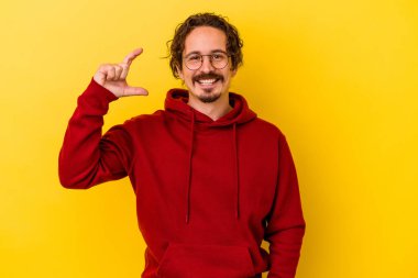 Sarı arka planda izole edilmiş genç beyaz adam işaret parmaklarıyla küçük bir şey tutuyor, gülümsüyor ve kendinden emin..