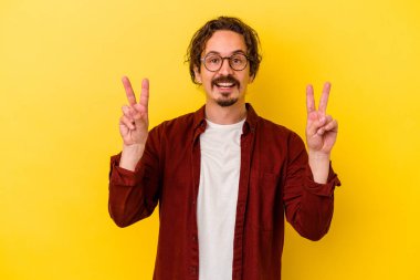 Sarı arka planda izole edilmiş genç beyaz adam zafer işareti gösteriyor ve gülümsüyor..