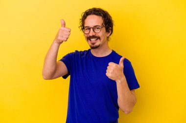 Sarı arka planda izole edilmiş genç beyaz adam iki parmağını da kaldırıyor, gülümsüyor ve kendinden emin..