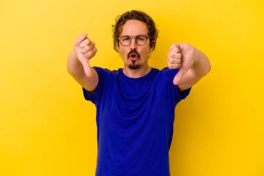 Sarı arka planda izole edilmiş genç beyaz adam baş parmağını aşağı gösteriyor ve hoşlanmadığını ifade ediyor..