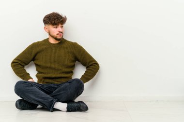 Faslı genç bir adam beyaz arka planda izole bir şekilde yerde oturuyor. Kafası karışmış, kendinden emin değil..