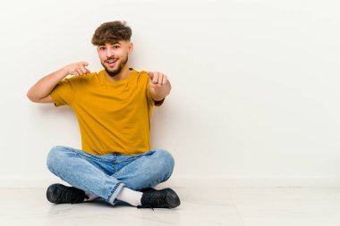 Faslı genç bir adam beyaz arka planda tek başına oturmuş, gülümsüyor ve öne bakıyor..