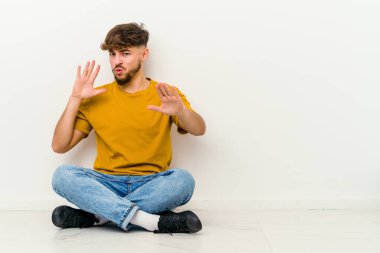 Genç Faslı bir adam beyaz arka planda izole bir şekilde yerde oturuyor. İğrenme hareketi gösteren birini reddediyor..