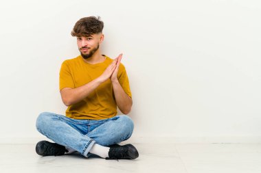 Genç Faslı bir adam beyaz arka planda izole bir şekilde yerde oturuyor enerjik ve rahat hissediyor, ellerini ovuşturuyor..