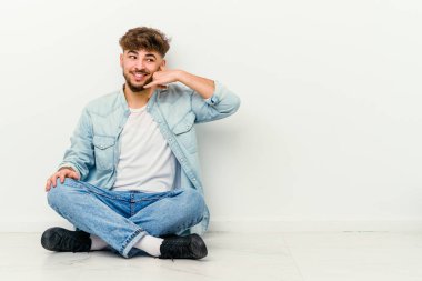 Faslı genç bir adam beyaz arka planda izole bir şekilde yerde oturuyor. Parmakları olan bir cep telefonu hareketi gösteriyor..