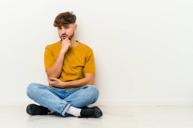 Faslı genç bir adam beyaz arka planda izole bir şekilde yerde oturuyor. Şüpheli, belirsiz, seni muayene ediyor..