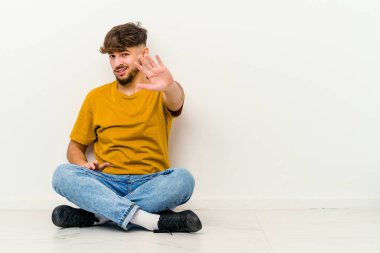 Faslı genç bir adam beyaz arka planda izole bir şekilde yerde oturuyor ve yakın bir tehlike nedeniyle şok oluyor.