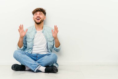 Faslı genç bir adam beyaz arka planda izole bir şekilde yerde oturuyor kahkahalar atıyor ve elini göğsüne koyuyor..