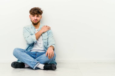 Beyaz arka planda izole bir şekilde yerde oturan Faslı genç bir adam omuz ağrısı çekiyor..