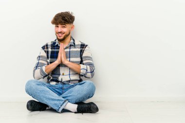 Faslı genç bir adam beyaz arka planda izole bir şekilde yerde oturuyor. El ele tutuşup dua ediyor. Kendine güveniyor..