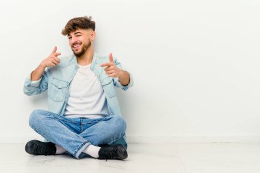 Faslı genç bir adam beyaz arka planda izole bir şekilde yerde oturuyor ve parmaklarıyla ön tarafı işaret ediyor..