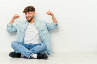 Faslı genç adam beyaz arka planda izole bir şekilde yerde oturuyor. Zaferden sonra yumruğunu kaldırıyor. Kazanan kavram..