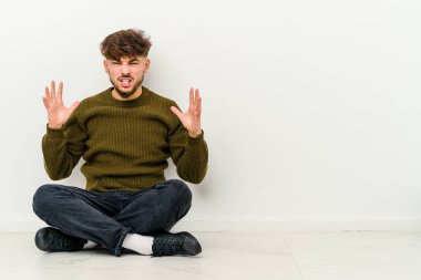 Faslı genç bir adam beyaz arka planda izole bir şekilde yerde oturmuş öfkeyle bağırıyor..