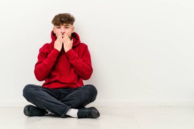 Faslı genç bir adam beyaz arka planda izole bir şekilde oturuyor. Şok olmuş, ağzı ellerle kaplanmış, yeni bir şey keşfetmek için sabırsızlanıyor..