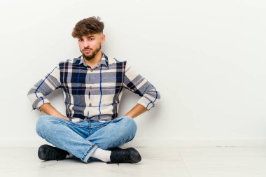 Faslı genç bir adam beyaz arka planda izole bir şekilde oturuyor hoşnutsuzca kaşlarını çatıyor, kollarını kavuşturuyor..