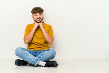 Beyaz arka planda izole bir şekilde yerde oturan genç Faslı adam ellerini çenesinin altında tutuyor ve mutlu bir şekilde yan tarafa bakıyor..