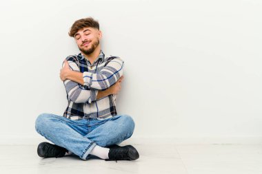 Genç Faslı bir adam beyaz arka planda izole bir şekilde yerde oturuyor, gülümsüyor ve mutlu..