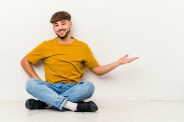 Beyaz arka planda izole bir şekilde yerde oturan Faslı genç adam hoş bir yüz ifadesi sergiliyor..