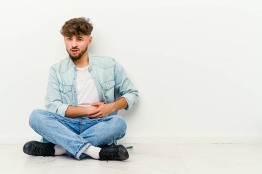 Genç Faslı bir adam beyaz arka planda karaciğer ve mide ağrısı şikayetiyle yerde yatıyor..