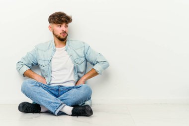 Faslı genç bir adam beyaz arka planda izole bir şekilde yerde oturuyor. Gülümsüyor, neşeli ve keyifli..