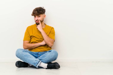 Faslı genç bir adam beyaz arka planda izole bir şekilde yerde oturuyor. İğneleyici bir ifadeyle kameraya bakıyor..