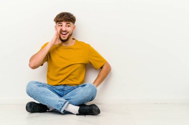 Faslı genç bir adam beyaz arka planda izole bir şekilde yerde oturmuş heyecanlı bir şekilde bağırıyor..