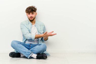 Faslı genç bir adam, beyaz arka planda izole bir şekilde yerde oturuyor. Elinde palmiye ağacının üzerinde bir kopyası var..