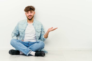 Faslı genç bir adam beyaz arka planda izole bir şekilde yerde oturuyor. Bir avuç içinde fotokopi alanı ve belinde başka bir el tutuyor..