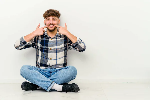 Νεαρός Μαροκινός Κάθεται Στο Πάτωμα Απομονωμένος Λευκό Φόντο Χαμογελά Δείχνοντας — Φωτογραφία Αρχείου