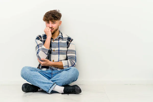 年轻的摩洛哥男子坐在地板上 被白色的背景隔离 咬着指甲 紧张而焦虑 — 图库照片