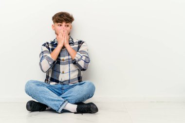 Genç Faslı bir adam beyaz arka planda izole bir şekilde yerde oturuyor. Düşünceli bir şekilde bir fotokopi odasına bakıyor. Ağzını eliyle kapatıyor..