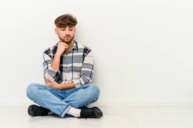 Faslı genç bir adam beyaz arka planda izole bir şekilde yerde oturuyor. Şüpheli ve şüpheci bir ifadeyle yana bakıyor..