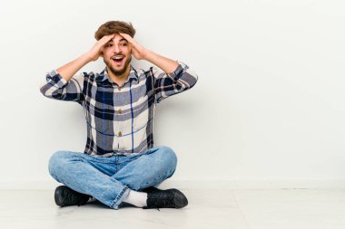 Beyaz arka planda izole bir şekilde yerde oturan genç Faslı adam ellerini başının üstünde tutarak neşeyle gülüyor. Mutluluk kavramı.