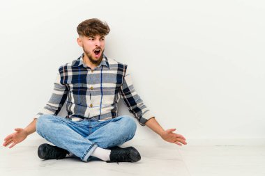 Genç Faslı bir adam beyaz arka planda izole bir şekilde yerde oturmuş kızgın, öfkeli ve hayal kırıklığına uğramış bir şekilde bağırıyor..