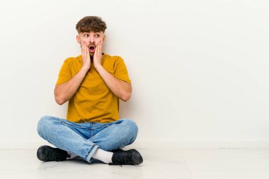 Genç Faslı bir adam beyaz arka planda yalnız başına korkmuş ve korkmuş bir şekilde yerde oturuyor..