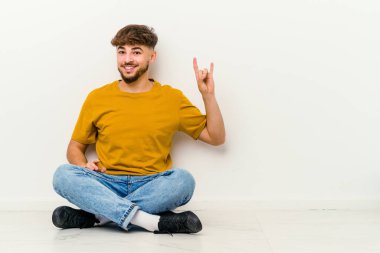 Beyaz arka planda izole bir şekilde yerde oturan genç Faslı bir adam devrim konsepti olarak boynuzlarını gösteriyor..