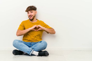 Faslı genç bir adam beyaz arka planda izole bir şekilde yerde oturuyor gülümsüyor ve elleriyle kalp şeklini gösteriyor..