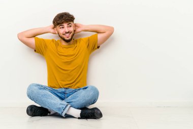 Faslı genç bir adam beyaz arka planda izole bir şekilde yerde oturuyor. Kendine güveniyor, elleri başının arkasında..